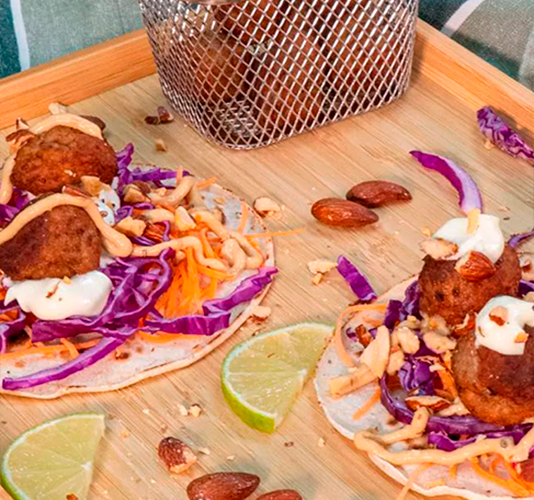 Tacos de albóndigas servidos en un plato de madera, cubiertos con col morada picada, zanahoria rallada, salsa blanca, almendras tostadas y perejil, acompañados de gajos de lima y una cesta metálica con más albóndigas.