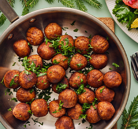 Albóndigas de carne o vegetales cocinadas en una sartén de acero inoxidable, espolvoreadas con perejil fresco picado, sobre un fondo verde y con un plato de ensalada al lado.