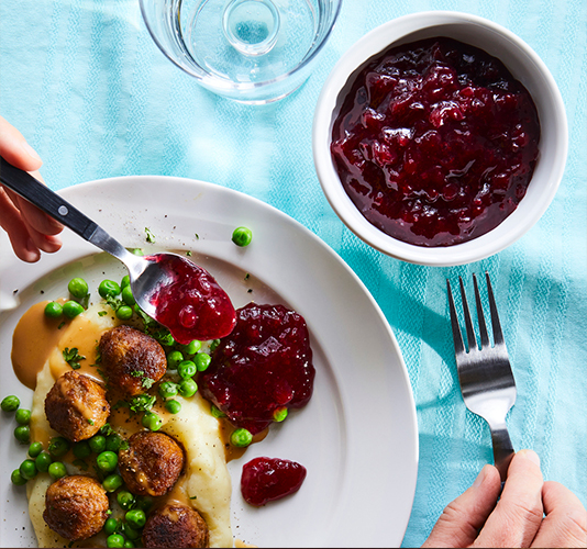 Plato blanco con puré de patatas, albóndigas y guisantes, acompañado de salsa de arándanos rojos y una copa de agua, con una persona sirviendo más salsa sobre el plato.