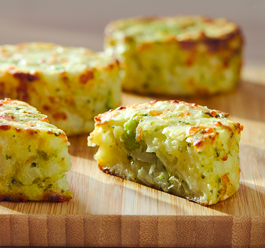 Medallones o tortitas de patata y verduras horneados con queso gratinado en la parte superior, presentados en una tabla de madera, con uno de ellos cortado para mostrar el interior.
