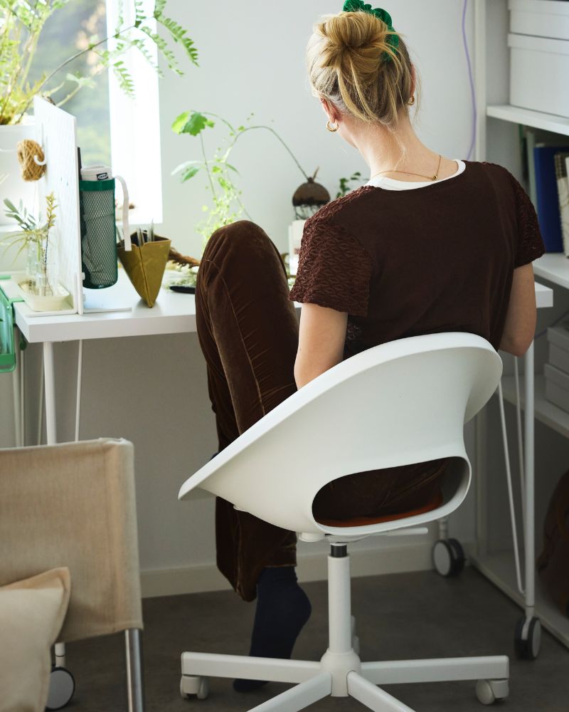 A person dressed in brown sitting on a white LOBERGET swivel chair by a white desk with green plants by a window.