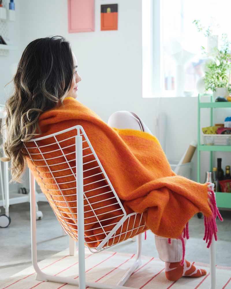 A person sitting in a white net chair wrapped in the orange KLIPPOXEL throw placed in a white room with colourful objects.