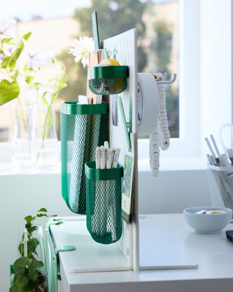 The free-standing SKÅDIS pegboard with green baskets and clips placed on the edge of a desk in front of a window.