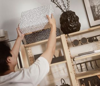 A person placing a RÄTTAREN storage box in white with black dots in an IVAR shelving unit in pine with black decoration.
