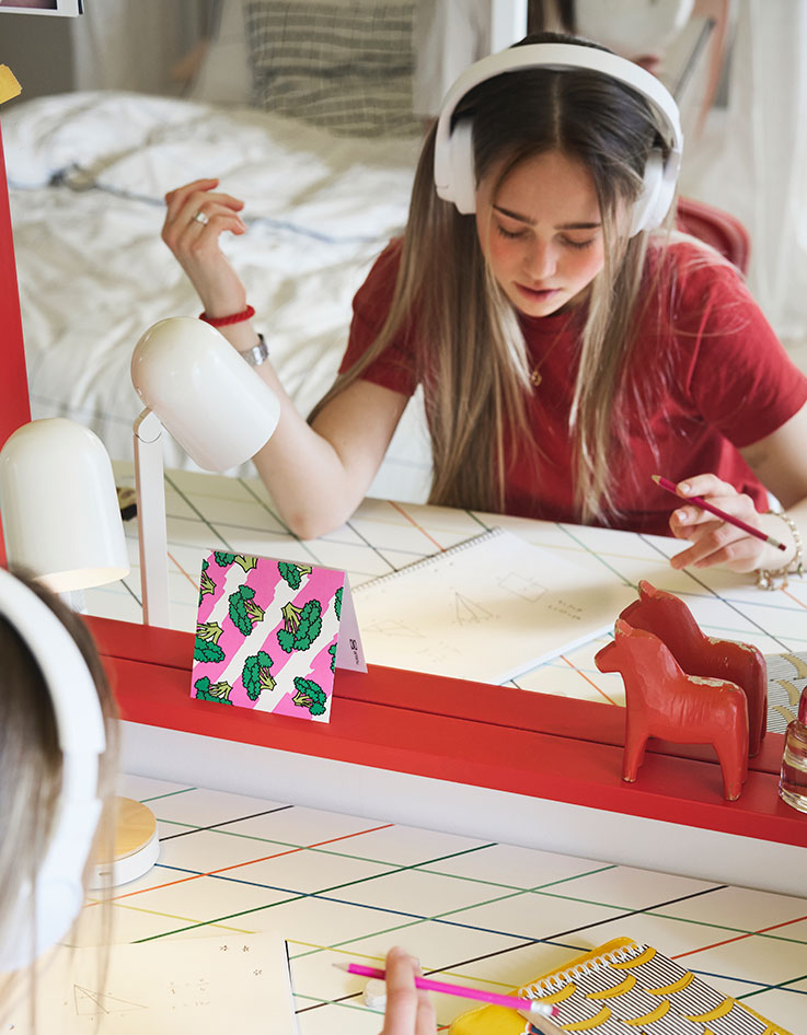  Un adolescente sentado estudiando junto a un escritorio LAGKAPTEN/ SPÄND con un colorido diseño cuadriculado colocado frente a un gran espejo rojo.