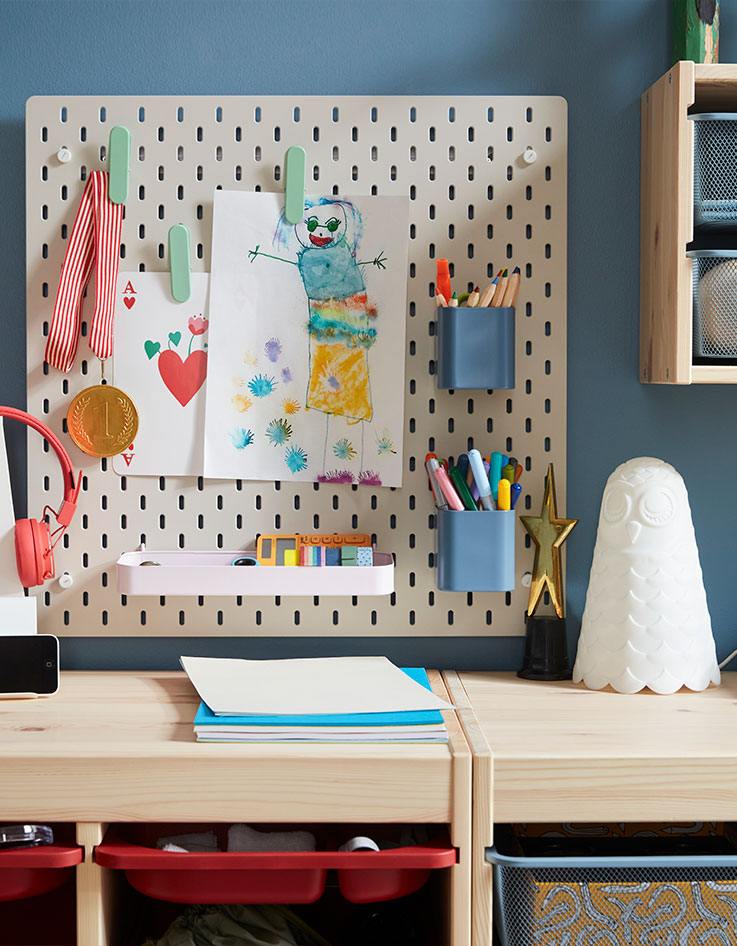 Un tablero de clavijas SKÅDIS de color beige con coloridos accesorios para sujetar bolígrafos y dibujos colocado en una pared azul sobre un armario de pino.