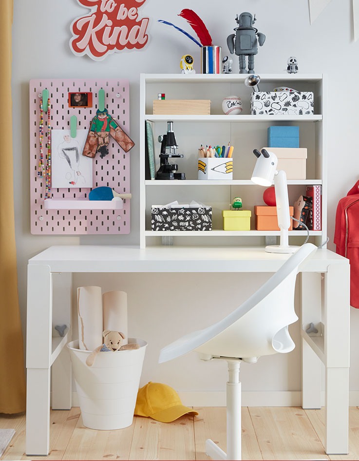 Un escritorio PÅHL blanco y una silla de escritorio blanca colocados en una habitación infantil con una estantería y un tablero de clavijas SKÅDIS rosa