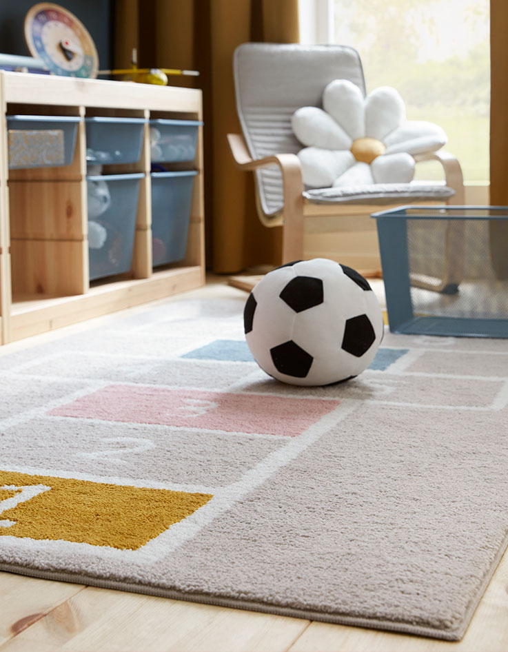 Una alfombra STÄPPSPARV con un motivo de rayuela colocada en una habitación infantil con un balón de fútbol textil y armarios de malla.
