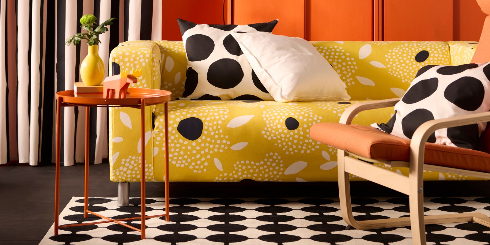 Sofá amarillo con cojines estampados blanco/negro. Mesa auxiliar naranja con jarrón amarillo. Alfombra estampada blanco/negro. Silla con cojín naranja.