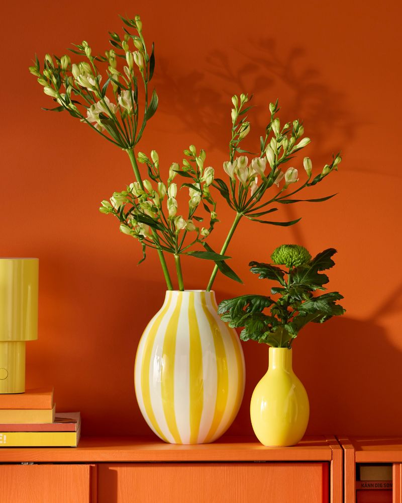 Dos jarrones amarillos, uno a rayas y otro liso, con flores blancas y plantas verdes sobre un gabinete naranja y pared naranja.