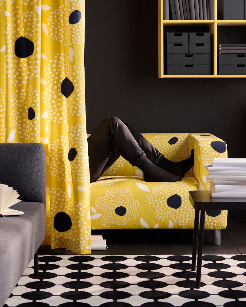 Las piernas de una persona son visibles en un sofá amarillo floral. Sofá gris, alfombra blanca y negra, y estantes amarillos con cajas negras.