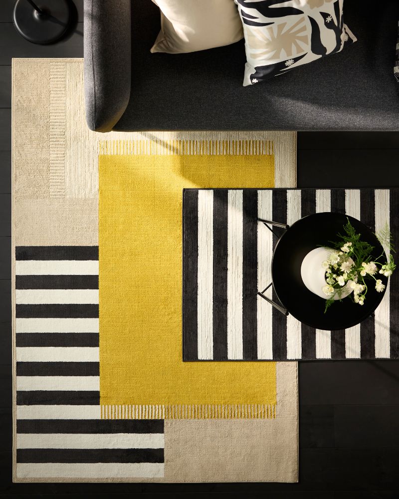 Vista aérea de un sofá negro con cojines estampados. Tres alfombras superpuestas (beige, amarilla, rayas blancas/negras) bajo una mesa de centro negra.