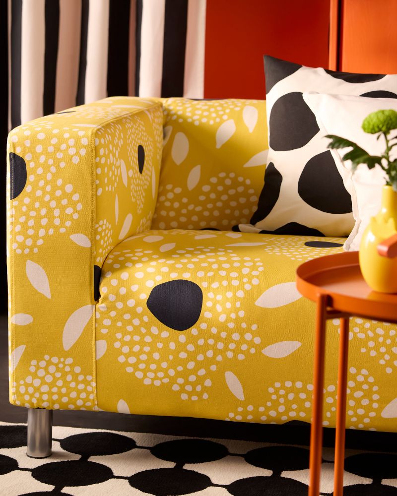 Un sofá moderno amarillo con patrones blancos y negros, un cojín blanco y negro. Pared roja y cortinas a rayas.
