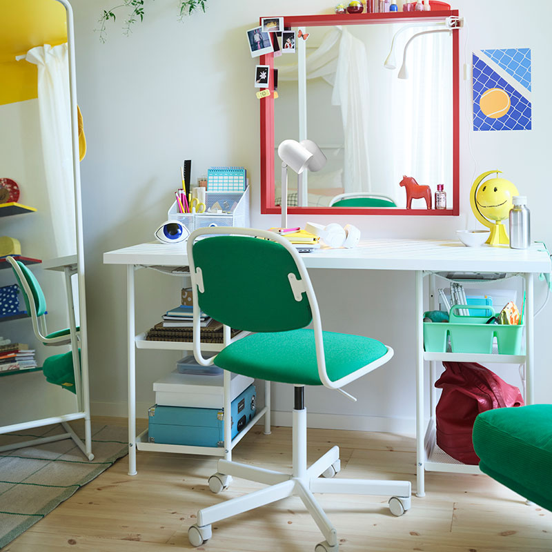 Un dormitorio juvenil con una gran cama blanca, un escritorio con un gran espejo rojo encima, una silla de escritorio y un sillón verdes, y mucho más.
