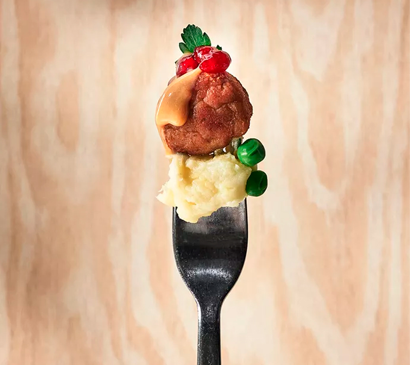 Una albóndiga de carne coronada con salsa, arándanos rojos y una ramita de perejil, apilada sobre un montículo de puré de patatas y dos guisantes verdes, todo ensartado en un tenedor.