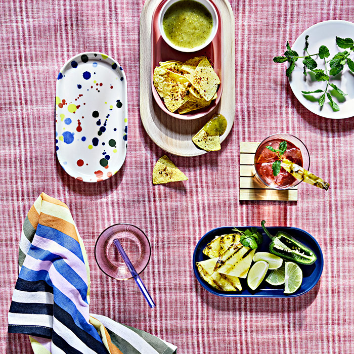 A summer table with a pink tablecloth. Bowls and plates with tortilla chips, green salsa, grilled pineapple, and a red drink.