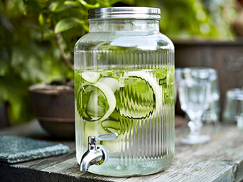 A glass beverage dispenser with water, cucumber slices, and a metal tap. It is placed on a wooden table.