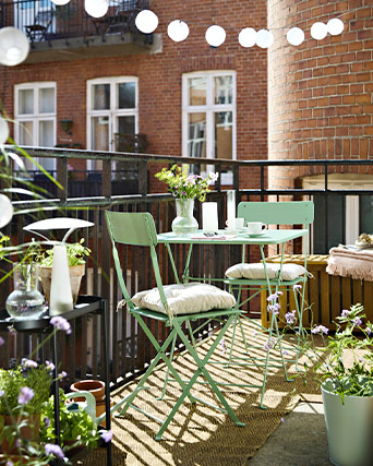A small balcony with a light green metal table and two folding chairs. There are flower pots and lights all around.