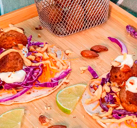 Tacos de albóndigas servidos en un plato de madera, cubiertos con col morada picada, zanahoria rallada, salsa blanca, almendras tostadas y perejil, acompañados de gajos de lima y una cesta metálica con más albóndigas.