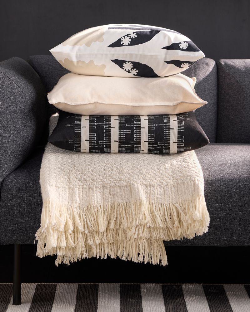 Primer plano de un sofá gris con una manta de flecos beige claro y una pila de tres cojines con patrones neutros y blanco/negro.
