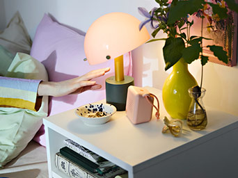Mesa de noche con lámpara rosa y jarrón amarillo. Una mano está a punto de apagar la luz, ideal para el momento de dormir.