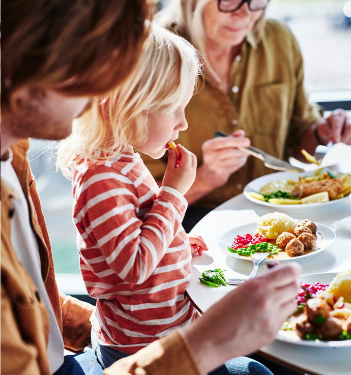 Dos adultos y un niño sentados en una mesa, disfrutando de una comida con platos que contienen albóndigas, puré de patatas, guisantes y arándanos, en un ambiente de restaurante o cafetería.