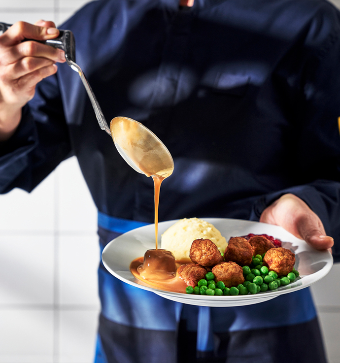 Persona sirviendo salsa oscura sobre un plato con albóndigas, puré de patatas, guisantes y salsa de arándanos rojos, creando una presentación clásica de comida sueca.