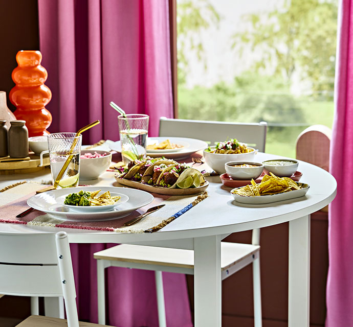 Mesa de comedor redonda blanca con platos de tacos, ensalada, nachos y cuencos pequeños. Cortinas magenta de fondo.