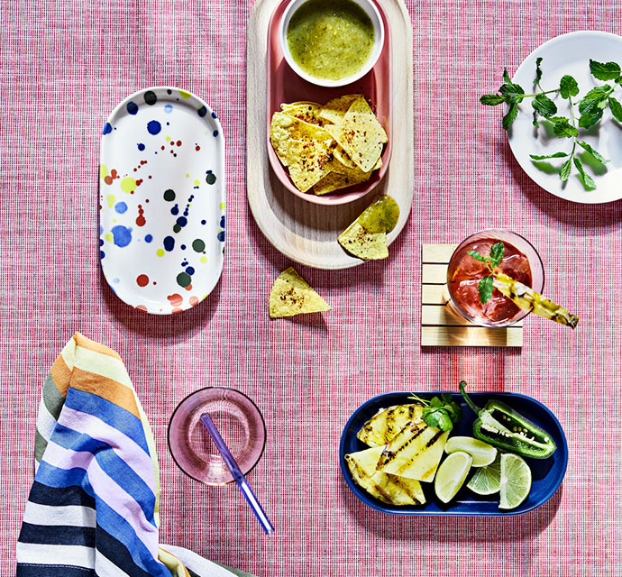 Comida y bebida en una mesa. Bol de nachos con salsa verde, plato de piña asada, limas y un cóctel.