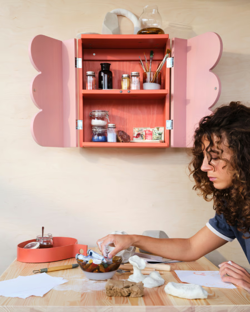Armario pequeño y rustico con pinceles y pinturas; chica pintando en mesa con cuencos y masas para esculturas
