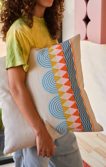 Persona con un cojín grande bajo el brazo, de colores otoñales, con formas geométricas planas y figuras.