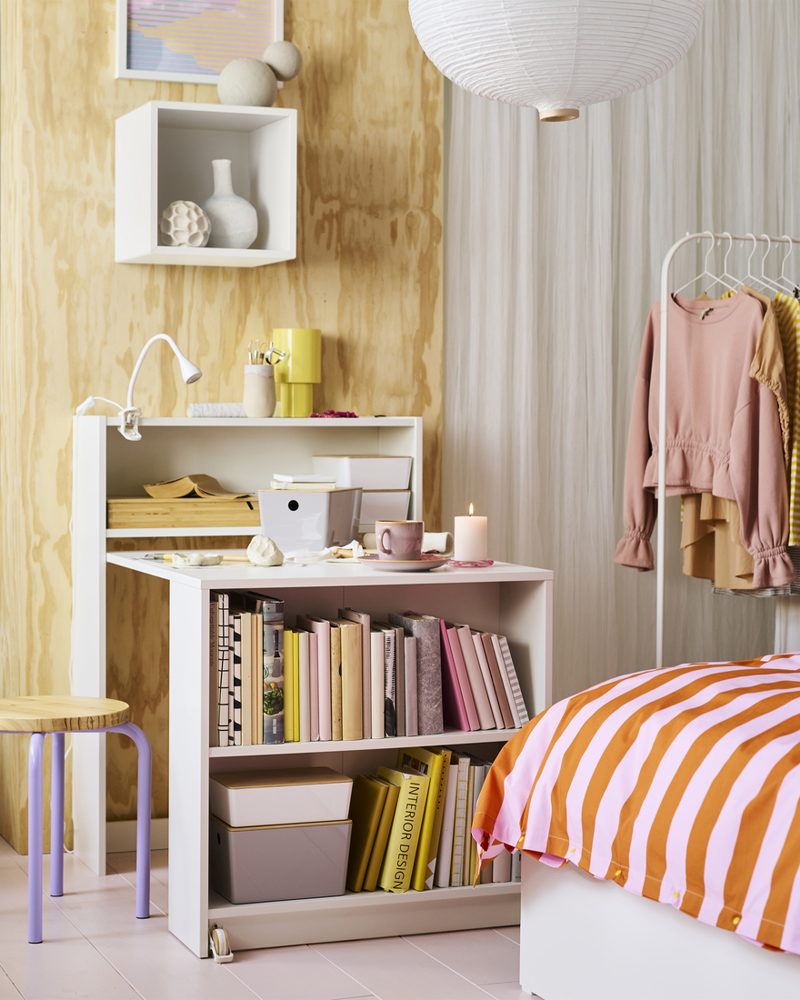 Un taburete DOMSTEN de color lila/pino junto a una estantería BILLY blanca con mesa plegable. La mesa tiene varios materiales de manualidades encima.