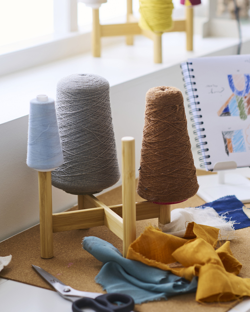 Un soporte para plantas DAKSJUS de bambú sobre un escritorio blanco con rollos de hilo de coser de diferentes colores y unas tijeras cerca.