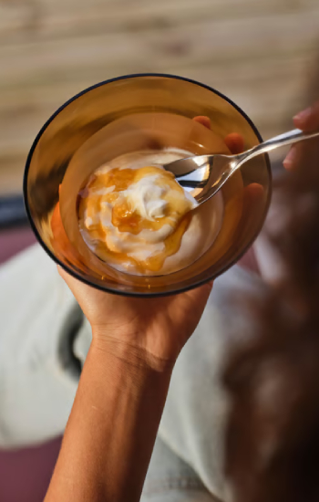 Mano sosteniendo un pequeño tazón de vidrio oscuro con yogur y miel.