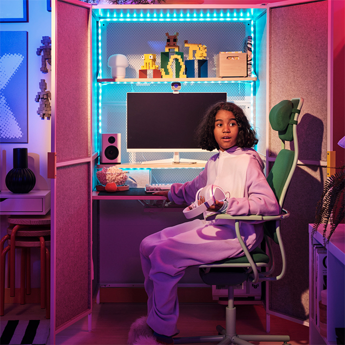Una joven sentada en una silla de oficina en un rincón de juego con luces de neón, vestida con un pijama lila, usando auriculares y mirando una pantalla de ordenador.
