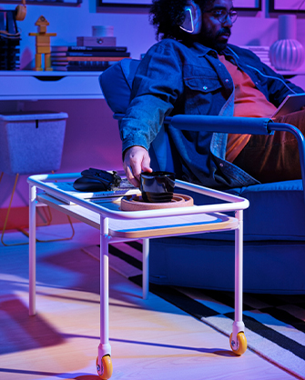 Un hombre sentado en un sillón azul, tomando una taza de café de una bandeja de servicio con ruedas de color blanco, en una habitación con iluminación de neón.