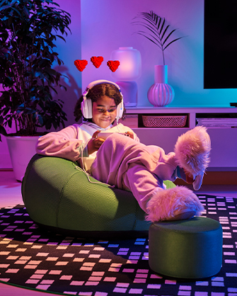 Una joven sentada en un puf de color verde en un salón con iluminación de neón, jugando con un dispositivo móvil mientras usa unos auriculares blancos. La escena está decorada con plantas y un puf pequeño a juego.