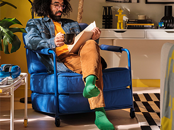 Un hombre con pelo rizado y barba, sentado en un sillón azul, leyendo un libro y tomando de una taza en una habitación con paredes en tonos amarillos y azules.