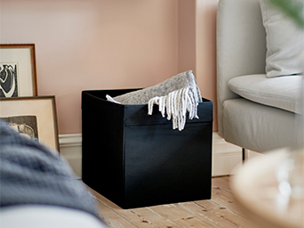 Black storage bin in a living room, with a gray blanket. Versatile solution for organizing blankets or cushions, perfect for the home.