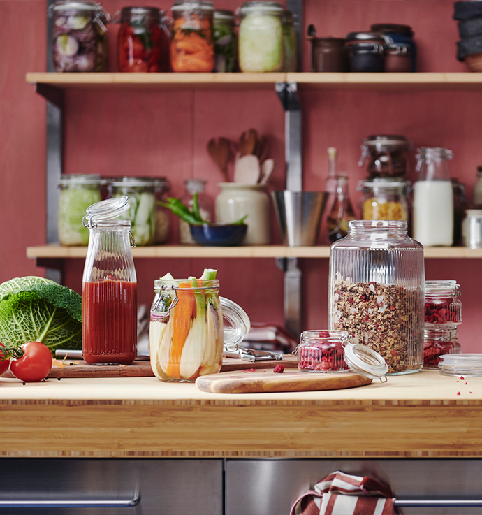 Encimera de cocina de madera con varios tarros de cristal. Se ven un tarro grande con granola, un tarro alto con zumo de tomate y otro con palitos de manzana y zanahoria. Detrás, en los estantes, hay más tarros llenos de conservas.