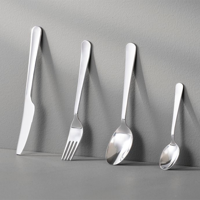 Un set de cubiertos de acero inoxidable con un acabado mate, compuesto por un cuchillo, un tenedor, una cuchara y una cucharilla, apoyados verticalmente en una pared de color gris claro.