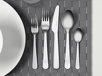 Un primer plano de un juego de cubiertos de acero inoxidable con el logo de la marca en la hoja del cuchillo. Están colocados sobre un mantel individual de tejido gris, junto a un plato blanco.