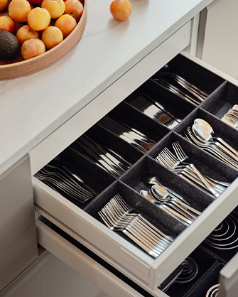 Cajón de cocina abierto con un organizador de fieltro gris, mostrando varios sets de cubiertos de acero inoxidable perfectamente ordenados. A la izquierda, un cuenco de madera con frutas.