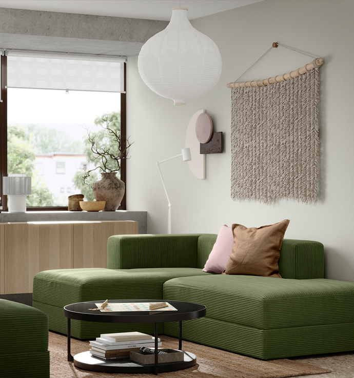 Green modular sofa with a large ceiling lamp, a round coffee table, and a textile wall decoration. The room has large windows looking outside.