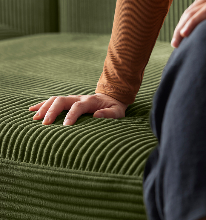 A close-up of a hand pressing the seat of an IKEA green modular sofa. The corduroy fabric with a ribbed texture is visible.