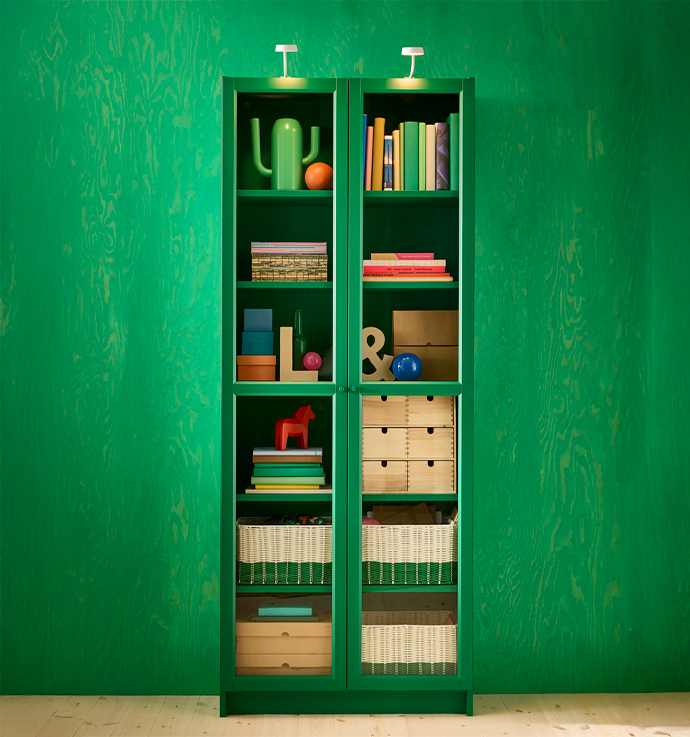  Un armario o estantería de color verde con puertas de cristal en un salón con papel pintado verde. El armario está lleno de libros, cajas de almacenaje y objetos decorativos como un cactus de cerámica.