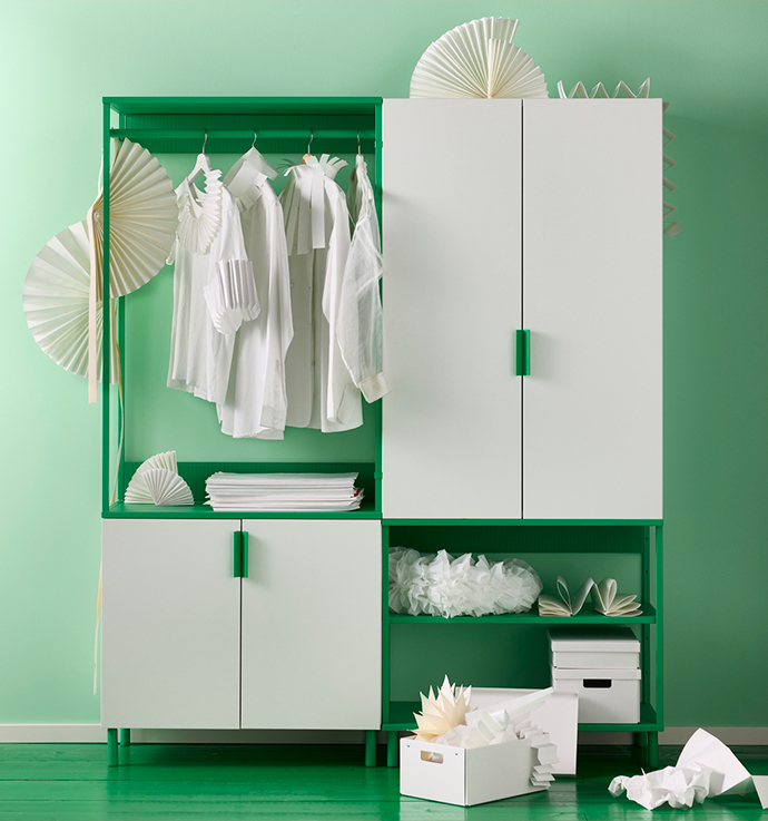 Un armario o solución de almacenaje verde y blanca, con un módulo de armario, otro con perchero y varios estantes. Dentro del armario y sobre las baldas se ve ropa y cajas. El fondo es una pared y un suelo pintados de verde.