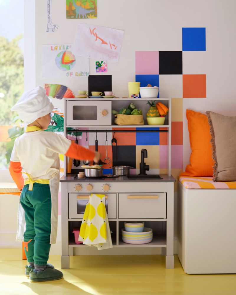 A child wearing a chef’s hat and an apron is standing by a grey SILLTRUT kitchen for children, stirring a pot on the hob. 