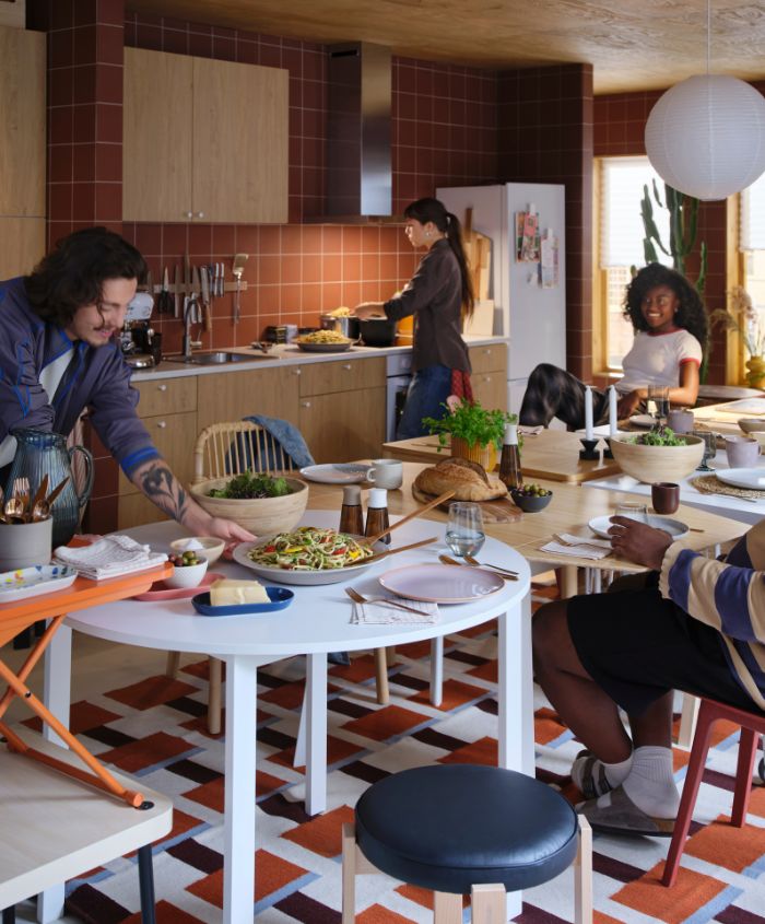 Four people are preparing for a meal in a lively kitchen. One of them is placing food on a set, white VIHALS table. 