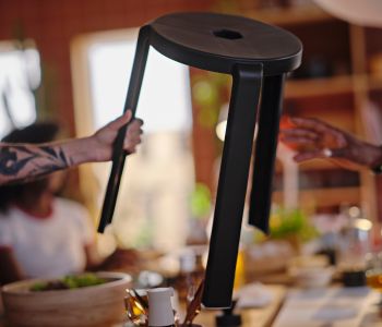 A tattooed arm is holding a dark grey IBBARBO stool in the air, reaching it over to a hand about to grab it. 
